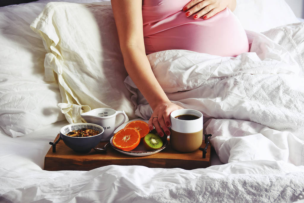 Schwangere Frau, die im Bett frühstückt - mit Obst und Kaffee