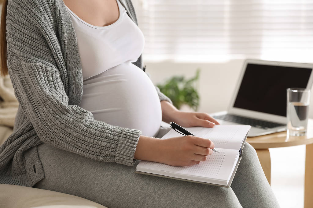 Schwangere Frau mit Notizbuch in der Hand, im Hintergrund ist Arbeitsplatz mit Notebook